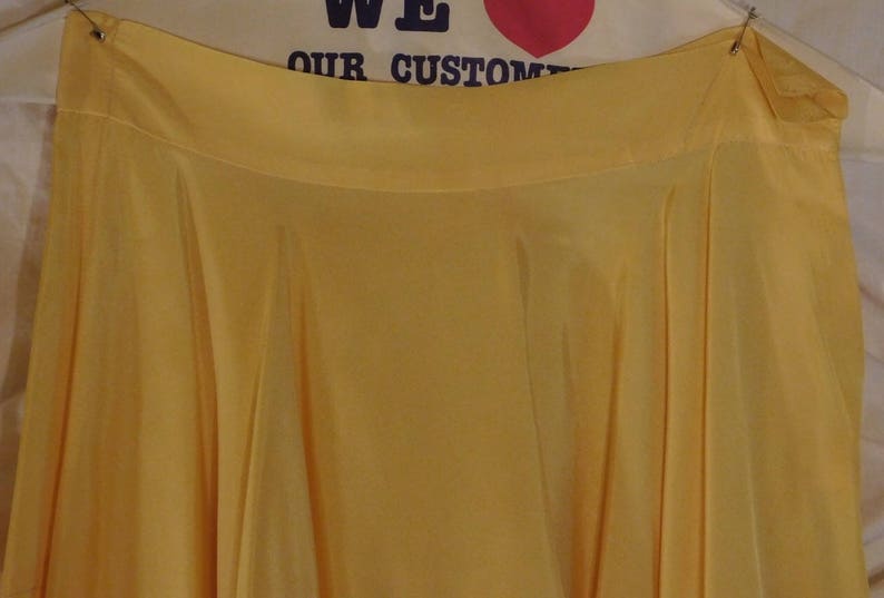 May include: A flowing, yellow skirt with a wide waistband. The fabric drapes in soft folds, creating a full silhouette. The skirt is suspended from a metal clip. The background includes the text "WE OUR CUSTOM".