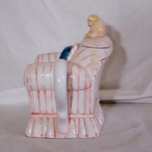 May include: A whimsical ceramic teapot shaped like an armchair. The teapot is white with red vertical stripes and a pink base. A small beige figurine sits on the lid. The handle is white, and a blue cushion is visible.