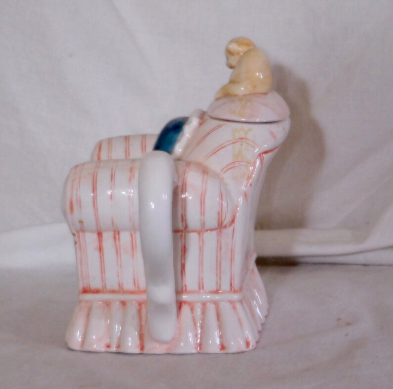 May include: A whimsical ceramic teapot shaped like an armchair. The teapot is white with red vertical stripes and a pink base. A small beige figurine sits on the lid. The handle is white, and a blue cushion is visible.