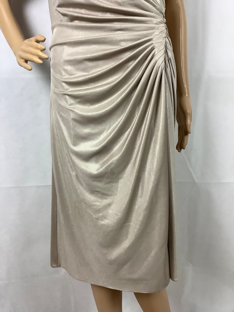 May include: A champagne-colored dress with a draped bodice and a knee-length skirt. The fabric has a subtle sheen, and the dress features a ruched detail on the side, adding texture and visual interest. The dress is displayed on a mannequin.