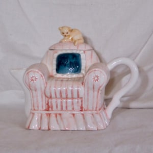 May include: A ceramic teapot shaped like a pink armchair with red stripes, a white handle, and a small beige cat figurine on top. The chair has a blue square with a floral design.