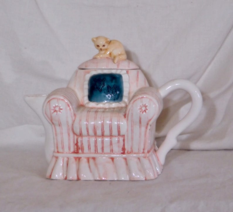 May include: A ceramic teapot shaped like a pink armchair with red stripes, a white handle, and a small beige cat figurine on top. The chair has a blue square with a floral design.