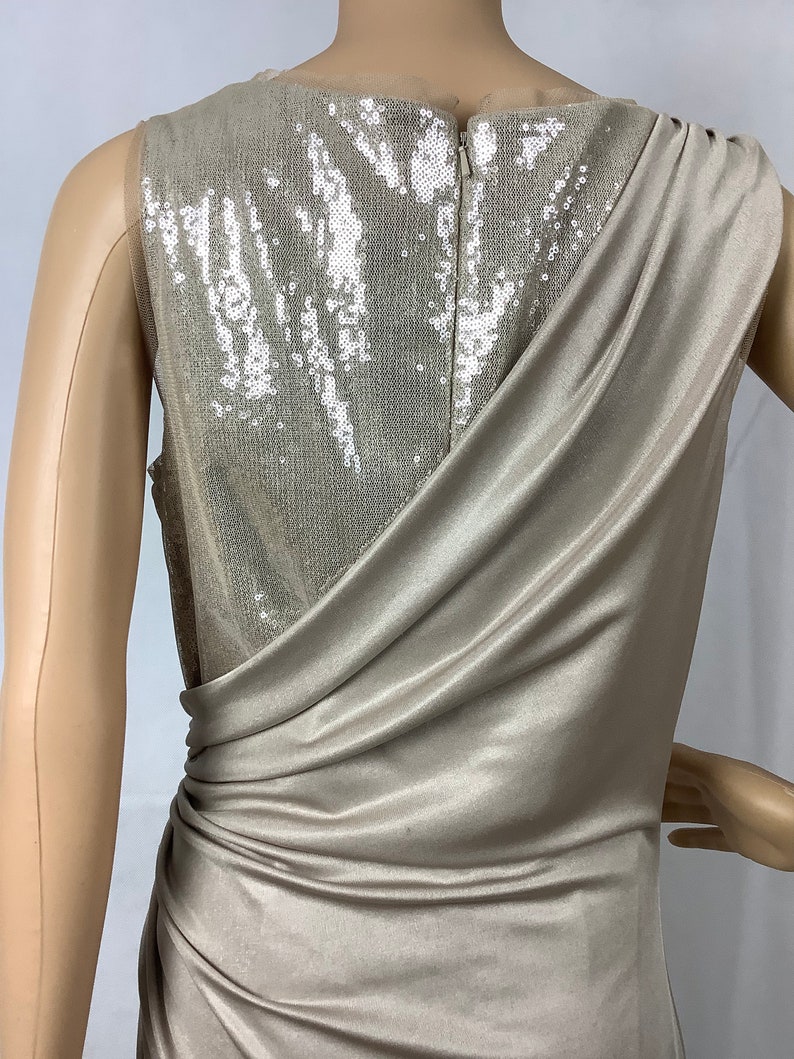May include: A champagne-colored formal dress with a draped, asymmetrical design. The back of the dress features a sequined panel, and a zipper closure. The dress is sleeveless and appears to be made of a soft, flowing fabric.