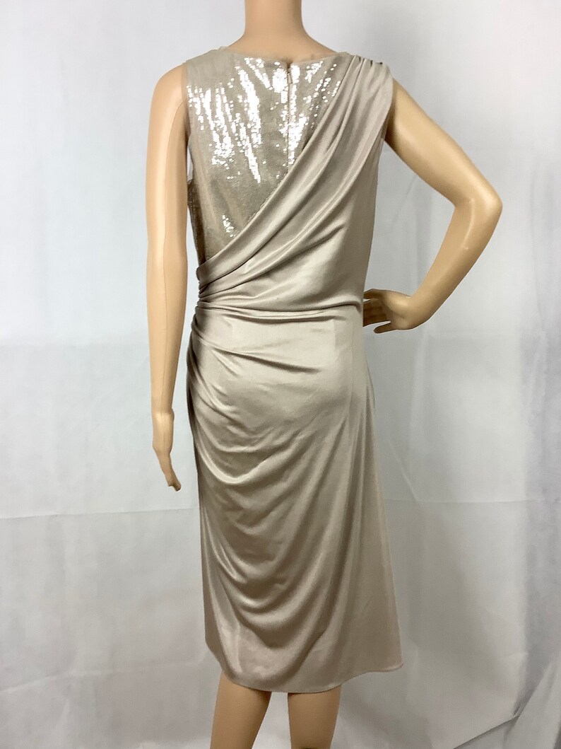 May include: A beige, sleeveless dress with a draped design. The back of the dress features a sequined panel. The dress has a knee-length hemline and is displayed on a mannequin.