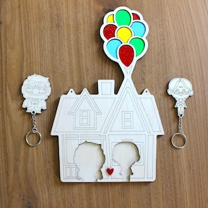 Könnte beinhalten: Holz-Schlüsselanhänger und Wanddekoration mit einem Haus mit einer Ballongruppe und Charakter-Ausschnitten. Die Ballons sind rot, gelb, grün und blau. Das Haus hat zwei Charaktersilhouetten und ein kleines rotes Herz.
