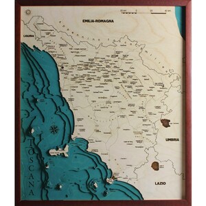 May include: A framed wooden map of the Tuscany region of Italy. The map is laser-cut with a detailed relief of the land and sea, with the sea in shades of teal and the land in natural wood tones. The frame is a dark reddish-brown.