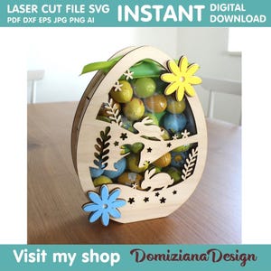 Può includere: Una scatola di legno a forma di uovo di Pasqua con un design a forma di coniglietti e fiori. La scatola è piena di caramelle colorate e ha un nastro verde attaccato alla parte superiore.