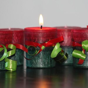 May include: Four red and green candles with a festive ribbon and green curly ribbon decoration. The candles are arranged in a row on a black surface.