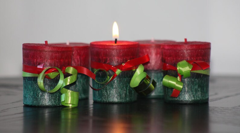 May include: Four red and green candles with a festive ribbon and green curly ribbon decoration. The candles are arranged in a row on a black surface.