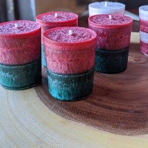 May include: Five red and green layered candles on a wooden surface. The candles are in a cylindrical shape and have a white wick.