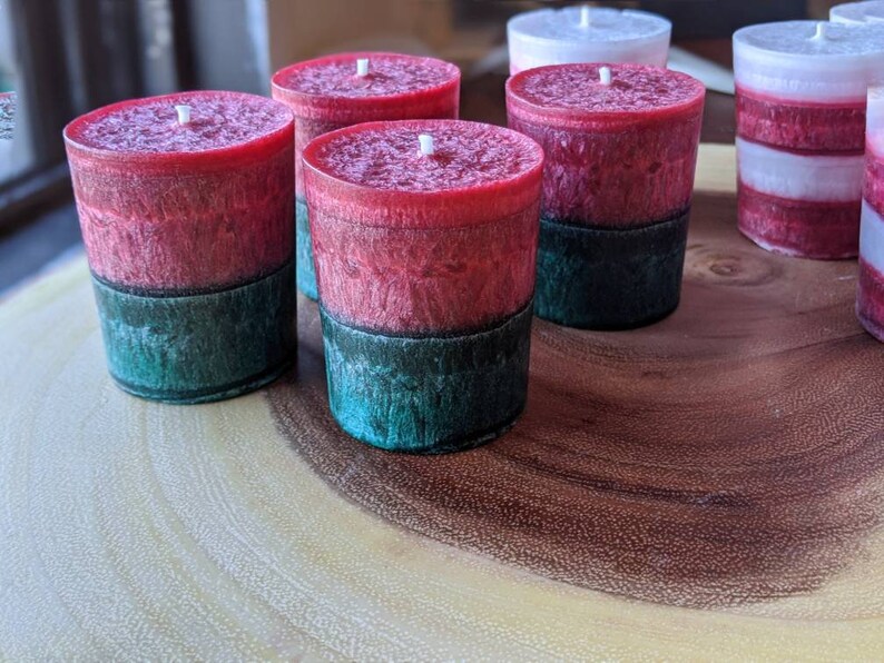 May include: Five red and green layered candles on a wooden surface. The candles are in a cylindrical shape and have a white wick.