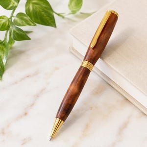 May include: A ballpoint pen crafted from polished dark brown wood with a prominent grain pattern. The pen features shiny gold-toned metal accents, including a slender pocket clip, a decorative ribbed center band, and a pointed tip at the base.