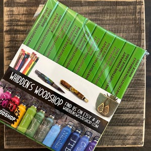 Pu&ograve; includere: Una collezione di borracce in acciaio inox personalizzate e colorate con diversi nomi. Le borracce sono disposte in fila su una superficie bianca con un cartello verde e dorato che recita "WHIDDEN'S WOODSHOP FIND US ON ETSY & AT WHIDDENSWOODSHOP.COM".