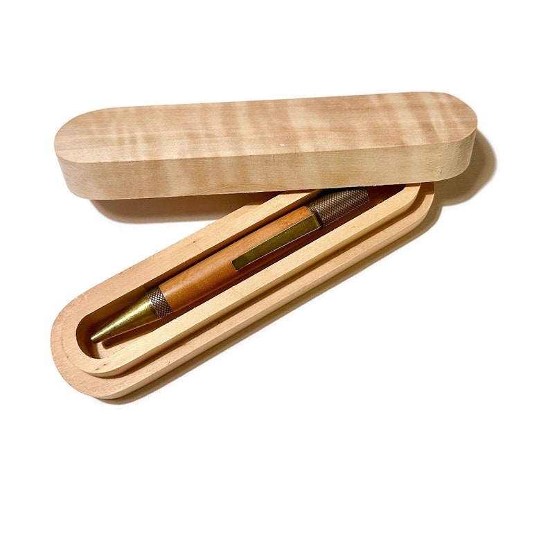May include: A wooden pen case with a lid, made of light wood with a natural grain pattern. The case is open and a wooden pen with a brass tip is inside.