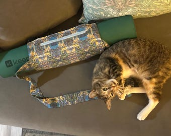 May include: A dark green yoga mat rolled inside a patterned fabric carrier bag featuring a colorful, intricate floral and scroll design with a white zipper. A brown and black tabby cat is lying next to the yoga mat carrier on a brown surface.