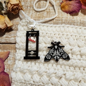 May include: Two black acrylic earrings, one shaped like a guillotine with a red blade and the other shaped like a moth with a skull and crossbones design.