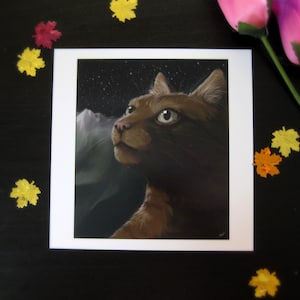 May include: A brown cat portrait print with a white border. The cat gazes upward against a starry night sky and a mountain range. Decorative leaves and pink flowers surround the print on a black surface.