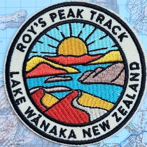 May include: A circular embroidered patch featuring a landscape of Roy's Peak Track, Lake Wanaka, New Zealand. The design shows a yellow sun, blue water, and red and brown mountains. The border text reads ROY'S PEAK TRACK LAKE WANAKA NEW ZEALAND.