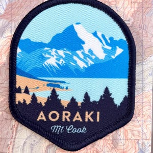 May include: A circular iron-on patch featuring a blue and white mountain landscape with a dark blue border. The text AORAKI Mt Cook is printed in white and gold. The patch is attached to a rectangular backing card with a topographic map design.