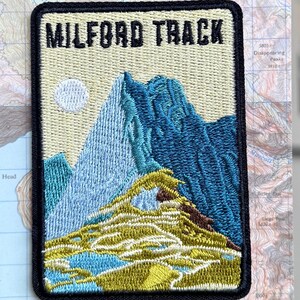 Op de afbeelding: Een rechthoekige geborduurde strijkpatch met een berglandschap met blauwe en groene toppen, een bleke zon en de tekst MILFORD TRACK bovenaan. De patch is bevestigd op een kaart met de tekst MILFORD TRACK Iron-on patch onderaan.