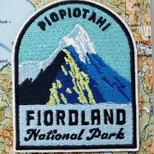 May include: An embroidered iron-on patch featuring a mountain landscape with light blue, dark blue, and yellow-green thread. The text reads PIOPOTAHI and FIORLAND National Park. The patch is mounted on a card with a map background and the text FIORLAND NATIONAL PARK Iron-on patch.