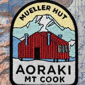May include: An embroidered iron-on patch featuring a red cabin with a black door and windows, set against a light blue mountain range and a pale yellow sky. The text reads MUELLER HUT at the top and AORAKI MT COOK at the bottom in black lettering.