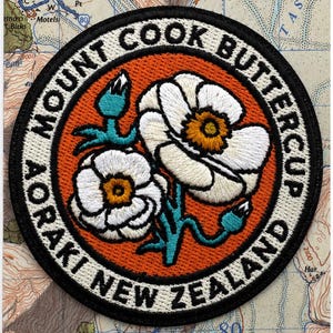 May include: A circular embroidered iron-on patch featuring two white buttercup flowers with yellow centers and teal stems on an orange background. The border text reads MOUNT COOK BUTTERCUP AORAKI NEW ZEALAND. The packaging text reads MOUNT COOK BUTTERCUP Iron-on patch.