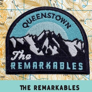 May include: A semi-circular embroidered patch featuring a mountain range in white and dark blue, with the text Queenstown and The Remarkables. The patch is mounted on a card with a topographic map background and the text The Remarkables Iron-on patch.