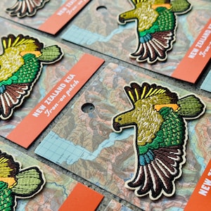Puede incluir: Parches bordados termoadhesivos con un diseño de loro Kea colorido. Los parches son en tonos verdes, amarillos y marrones, con patrones detallados de alas y plumas. Cada parche se muestra sobre un fondo similar a un mapa con una etiqueta naranja que dice "NEW ZEALAND KEA Iron-on patch."