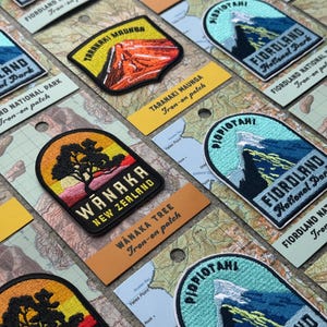 Könnte beinhalten: Gestickte Aufnäher zum Aufbügeln mit Landschaftsmotiven aus den Nationalparks Neuseelands. Die Aufnäher zeigen farbenfrohe Designs von Bergen und Bäumen, mit Texten wie "Wanaka" und "Fiordland National Park".
