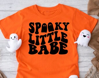 Könnte beinhalten: Orangefarbenes T-Shirt mit kurzen Ärmeln und schwarzem Text, der "Spooky Little Babe" in einer Retro-Schriftart lautet. Der Text ist von drei schwarzen Geister-Silhouetten umgeben.