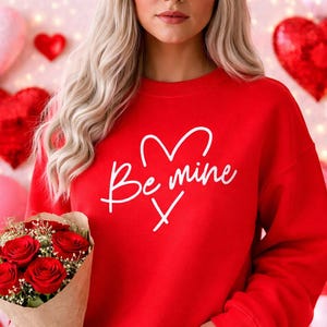以下が含まれることがあります： 白い筆記体で「Be mine」とハートのデザインが施された赤いスウェットシャツ。茶色の紙に包まれた赤いバラの花束を持っています。背景にはハート型の装飾が施されています。