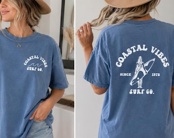 May include: A blue short-sleeved t-shirt with the text "Coastal Vibes Surf Co." on the front and back. The back of the shirt features a graphic of a person with a surfboard. The shirt is made of a soft material.