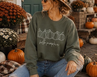Könnte beinhalten: Ein olivgrünes Sweatshirt mit Rundhalsausschnitt, das eine weiße Linienzeichnung von fünf Kürbissen und den kursiven Schriftzug "hello pumpkin" auf der Brust zeigt. Der langärmelige Pullover hat einen gerippten Ausschnitt, Bündchen und Saum.