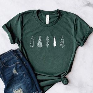 May include: Dark green t-shirt featuring a white line drawing of five different Christmas trees. The shirt is paired with blue denim shorts. The t-shirt is made of a soft material.