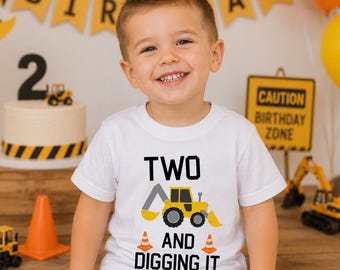May include: A young child wearing a white t-shirt with a yellow construction vehicle graphic and the text "TWO AND DIGGING IT." The child is smiling in front of a birthday banner and cake.