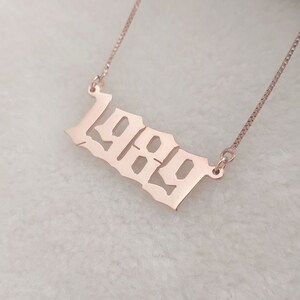 May include: A rose gold necklace with a pendant in the shape of the year 1989. The pendant is in a gothic font.
