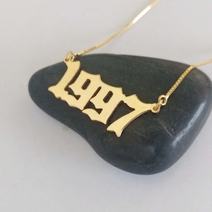 Puede incluir: Collar de oro con un colgante en forma del año 1997. El colgante está en una fuente de estilo gótico.