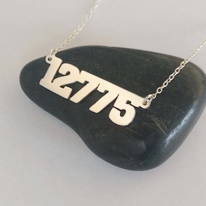 May include: Silver necklace with a pendant featuring the number "12775" in a bold, modern font.