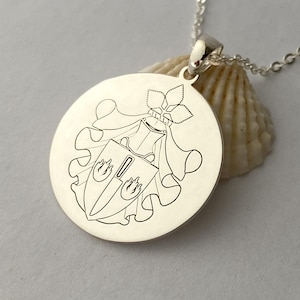 May include: A silver pendant necklace. The circular pendant features an engraved coat of arms design. The necklace hangs from a delicate chain. The background includes a seashell.