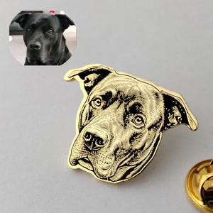 May include: A gold-toned metal lapel pin featuring a detailed black engraved illustration of a dog's head. The pin is shaped like the dog's face and includes a circular metal clutch backing. A small circular inset shows the original black dog.