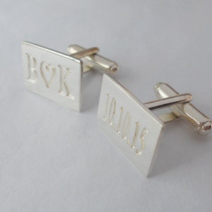 Puede incluir: Gemelos de plata con un diseño rectangular. Un gemelo está grabado con las iniciales "R & K" con un símbolo de corazón entre las letras. El otro gemelo está grabado con la fecha "10.10.15".