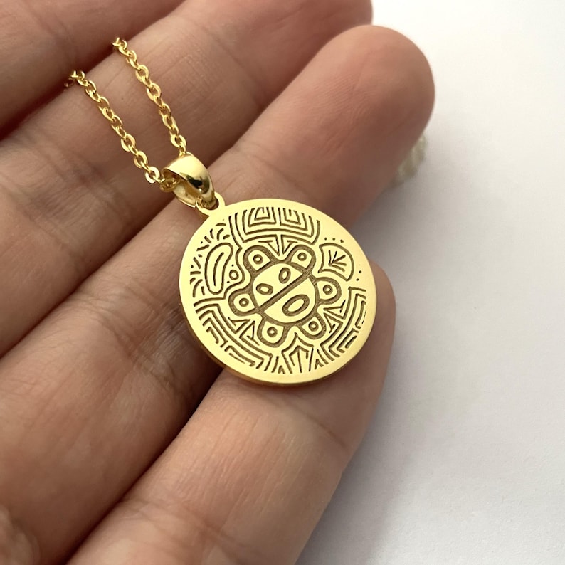 May include: A gold-colored pendant necklace featuring an engraved circular pendant with an intricate design. The pendant hangs from a delicate chain. The design on the pendant is a series of lines and shapes.