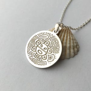 May include: A circular silver pendant featuring an engraved stylized sun-like figure with geometric patterns, suspended from a delicate silver chain. The pendant rests against a textured, off-white seashell, highlighting the metallic finish.