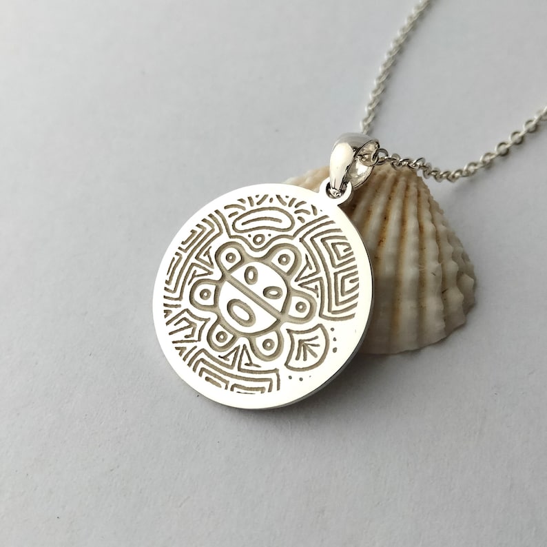 May include: A circular silver pendant featuring an engraved stylized sun-like figure with geometric patterns, suspended from a delicate silver chain. The pendant rests against a textured, off-white seashell, highlighting the metallic finish.