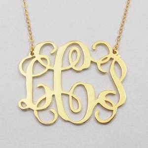 May include: A gold-toned necklace with a delicate chain and a large, open-work monogram pendant. The pendant features three intertwined letters in a cursive script.