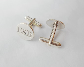 May include: A pair of silver cufflinks with a round, oval-shaped face. The face of the cufflink is engraved with the initials "ESB".