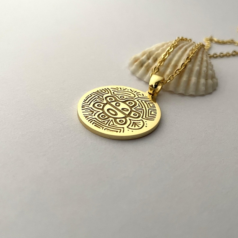 May include: Gold-colored pendant necklace with a circular pendant featuring an engraved design. The pendant hangs from a delicate chain. A seashell rests in the background. The necklace is displayed on a white surface.
