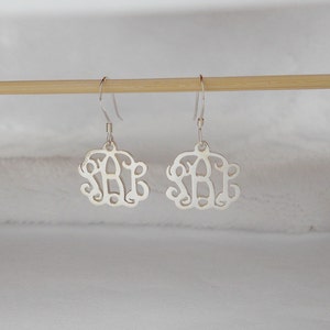 Könnte beinhalten: Ein Paar silberne Monogrammohrringe mit einem zarten, kursiv geschriebenen Design. Die Ohrringe hängen an einer Holzoberfläche.