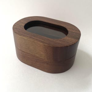 Puede incluir: Una caja de joyería de madera con un acabado marrón oscuro. La caja tiene forma ovalada con una ventana ovalada transparente en la parte superior. La caja está cerrada, con una costura visible donde se unen la tapa y la base.